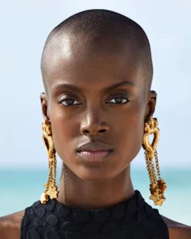 Madisin Rian poses in front of the ocean in a black onesie swimsuit and oversized gold earrings.